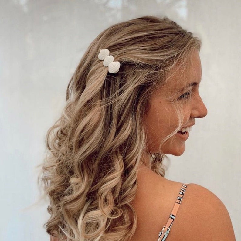 Pu&ograve; includere: Primo piano di una persona con capelli biondi ondulati, adornati con tre fermagli per capelli a forma di conchiglia bianca. Le clip sono raggruppate, aggiungendo un tocco balneare all'acconciatura. La persona sorride e indossa un top fantasia.