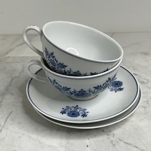 Op de afbeelding: Twee witte theekopjes en schotels met blauwe bloemmotieven. De kopjes zijn op elkaar gestapeld, elk met een oor. De schotels hebben een bijpassend blauw bloemmotief en een blauwe rand. De set staat op een wit oppervlak.