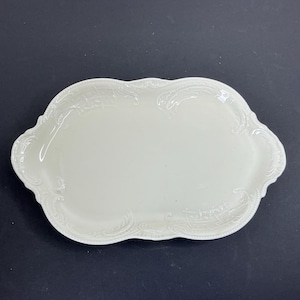 Op de afbeelding: Een ovaalvormige gebroken witte keramische serveerschaal met een glad centraal oppervlak en een decoratieve verhoogde reliëfrand met ingewikkelde krul- en filigraanpatronen langs de randen, wat zorgt voor een textuurrijk kader.