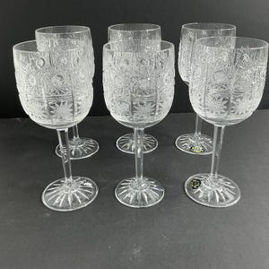 Op de afbeelding: Een set van zes wijnglazen van helder kristal met ingewikkelde geslepen patronen, waaronder sterren en geometrische motieven. Elk glas heeft een hoge, slanke steel en een ronde voet met een stralend sterpatroon in het glas gegraveerd.