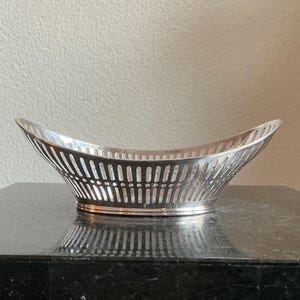 Könnte beinhalten: Ein silberfarbener, ovaler Korb mit durchbrochenem Design. Der Korb hat eine breite, offene Oberseite und einen leicht gebogenen Rand. Der Korb steht auf einer dunklen Oberfläche.