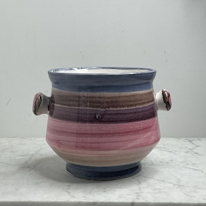 Op de afbeelding: Een keramische vaas met een afgeronde body en twee kleine handvatten. De vaas heeft horizontale kleurstroken, waaronder blauw, bruin, paars en roze. De vaas wordt waarschijnlijk gebruikt voor decoratieve doeleinden of om bloemen in te zetten.