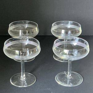 Op de afbeelding: Een set van vier heldere glazen coupes, elk voorzien van een reeks dunne, horizontale geëtste lijnen rondom de bovenste kom. Deze glazen op voet hebben een brede, ondiepe kelk en een ronde voet, met een consistent ontwerp voor alle vier.