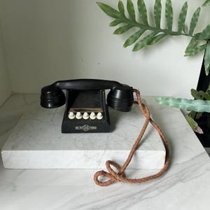 Op de afbeelding: Een antieke zwarte telefoon met een gebogen handset en een reeks witte knoppen. De telefoon heeft een getextureerd uiterlijk en een bruin, gevlochten snoer. De telefoon wordt gepresenteerd op een wit marmeren oppervlak.