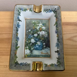 Op de afbeelding: Een rechthoekige porseleinen asbak met een centrale afbeelding van een blauw-wit bloemenboeket in een vaas. De rand is versierd met een delicaat blauw en groen bloemenrankpatroon, afgewerkt met een goudkleurige rand en twee goudkleurige inkepingen.