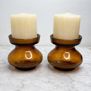 Op de afbeelding: Twee amberkleurige glazen kandelaars met een afgeronde basis en een uitlopende bovenkant. Elke houder ondersteunt een dikke, gebroken witte pilaar kaars. De kandelaars hebben een vintage esthetiek, geschikt voor woondecoratie.