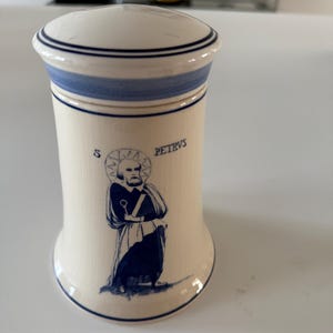 Op de afbeelding: Een witte keramische pot met een blauw-wit gestreept deksel. De pot heeft een blauwe en witte illustratie van een man met een halo en de tekst "S. PETRVS".