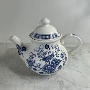Könnte beinhalten: Weiße Keramik-Teekanne mit Deckel, Ausguss und Henkel. Die Teekanne ist mit blauen Blumen- und Vogelmustern verziert. Der Deckel hat einen kleinen Knopf und ein geometrisches blaues Design. Die Teekanne steht auf einer Marmoroberfläche.