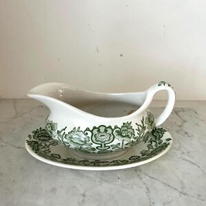 Pode incluir: Um molho branco com um design floral verde e um pires correspondente. O molho tem uma base larga e arredondada e um bico de um lado. O pires tem uma borda ondulada.