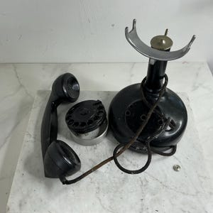 Op de afbeelding: Een antieke zwarte draaischijftelefoon, gedemonteerd. De componenten omvatten de basis, de handset, de draaischijf en een vork. De telefoon heeft een zilveren vork en een messing knop. De telefoon staat op een wit-grijs marmeren oppervlak.