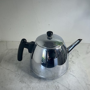 Op de afbeelding: Een glanzende, verchroomde theepot met een zwarte handgreep en knop. De theepot heeft een gebogen tuit en een afgeronde body. Het gepolijste oppervlak reflecteert de omgeving. Een klassiek keukenartikel.