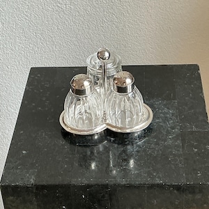 Puede incluir: Un juego de salero y pimentero vintage con un tarro de cristal a juego para especias. El juego incluye dos saleros de cristal transparente y acanalado con tapas de metal plateado y un tarro pequeño con tapa. El juego se encuentra sobre una bandeja de metal plateado.
