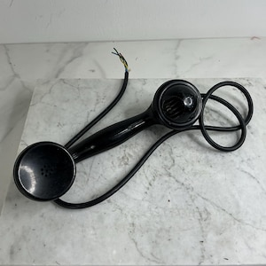 Può includere: Ricevitore telefonico nero vintage con auricolare e bocca circolari. Il ricevitore ha un'impugnatura curva e un cavo a spirale. I fili sono visibili all'estremità del cavo. Il telefono è appoggiato su una superficie di marmo.
