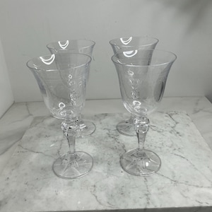 Può includere: Quattro calici da vino trasparenti con disegni floreali incisi. I calici hanno una coppa a forma di tulipano, uno stelo decorativo e una base circolare. Sono disposti su una superficie di marmo bianco e grigio.