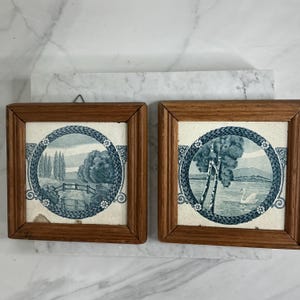 Può includere: Due piastrelle paesaggistiche incorniciate. Ogni piastrella presenta un design panoramico blu e bianco all'interno di un bordo circolare. Uno raffigura un ponte e alberi, l'altro un cigno su un lago. Le cornici sono in legno.
