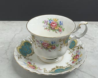 May include: A white porcelain teacup and saucer set featuring a scalloped edge, gold trim, and a colorful floral pattern of pink roses and blue flowers. The design includes ornate turquoise and gold scrollwork accents on the cup and saucer.