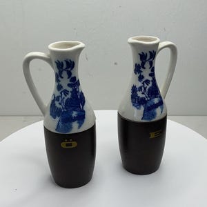 Op de afbeelding: Twee decoratieve keramische kruiken. Het bovenste gedeelte is wit met blauwe bloemmotieven en de onderkant is donkerbruin. Elke kruik heeft een wit handvat en een klein gouden embleem.
