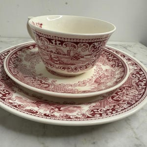 Könnte beinhalten: Ein cremefarbenes Vintage-Teeservice mit einer Tasse, Untertasse und passendem Teller. Das Set zeigt ein detailliertes bordeauxfarbenes Transferware-Design einer Dorflandschaft. Die Tasse hat einen Henkel und das Set ist auf einer Marmoroberfläche angeordnet.