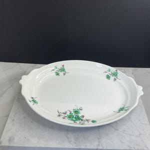 May include: An oval white ceramic serving platter featuring decorative handles on each side. The platter is adorned with small, scattered floral motifs in shades of green and grey, distributed across the smooth, glossy white surface.