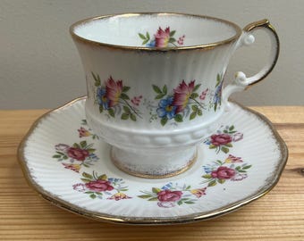 May include: A white porcelain teacup and saucer set featuring a delicate floral pattern of pink and blue flowers with green leaves. The rim of the cup and the edge of the saucer are trimmed with gold, and the cup has a decorative handle.