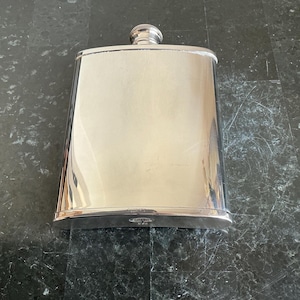 Può includere: Una fiaschetta rettangolare in acciaio inossidabile lucido con la parte superiore arrotondata e un tappo a vite. La fiaschetta ha una superficie lucida e riflettente e un piccolo tappo decorativo. La fiaschetta è progettata per contenere liquidi.