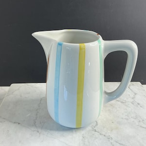 Może przedstawiać: Biały ceramiczny dzbanek ozdobiony pionowymi paskami w pastelowych odcieniach jasnego błękitu, żółci i miętowej zieleni. Dzbanek ma gładkie, błyszczące wykończenie, zakrzywiony uchwyt po prawej stronie oraz wyraźnie zaznaczony dzióbek po lewej.