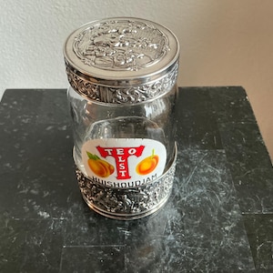 Peut inclure: Un bocal en verre transparent avec un couvercle et une base en métal argenté. Le couvercle est orné de motifs floraux et de volutes. Le bocal porte une étiquette avec le texte "TEO LST HuisHoudJam" et des images d'abricots.