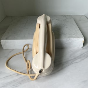 Può includere: Telefono vintage color crema con cornetta e cavo a spirale. Il telefono ha un design aerodinamico con una base arrotondata e un ricevitore lungo e curvo. Il cavo è di colore beige chiaro.