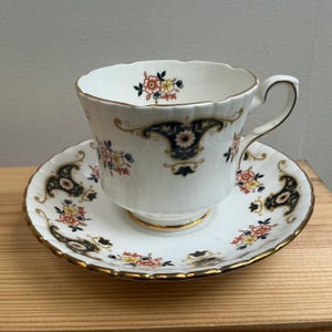 Op de afbeelding: Een wit porseleinen theekop en schotel met een geschulpte rand en gouden afwerking. Het ontwerp bevat donkerblauwe, sierlijke krullen en kleine trossen roze en gele bloemen. De kop heeft een gebogen handvat en een gouden rand aan de onderkant.