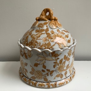 Op de afbeelding: Een witte keramische pot met een deksel, versierd met een bloemenpatroon in bruintinten. Het deksel heeft een decoratieve knop.