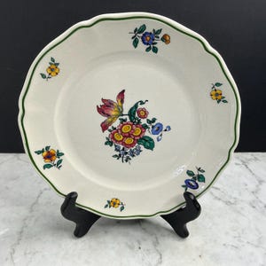Op de afbeelding: Een ronde keramische bord met een geschulpte rand en een dunne groene rand. Het midden is voorzien van een kleurrijk bloemenboeket met roze, rode en blauwe bloemen. Kleinere individuele bloemtakjes zijn verspreid over de rand van het bord.