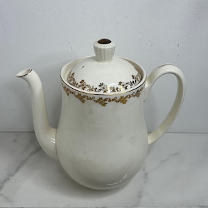 Può includere: Teiera in ceramica color crema con beccuccio e manico curvi. Il coperchio e il corpo sono adornati da un motivo floreale dorato. Il coperchio ha un pomello marrone. La teiera è su una superficie bianca.