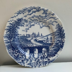 May include: Blue and white ceramic plate with a scenic design of a bridge over a river, a house in the distance, and a person fishing. The plate has a white background with blue details.