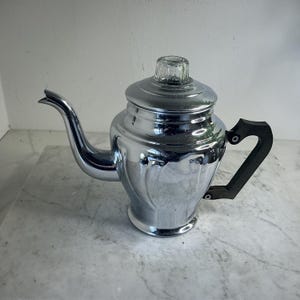 Op de afbeelding: Een vintage chromen theepot met een zwarte handgreep en een doorzichtige glazen knop op het deksel. De theepot heeft een gebogen tuit en staat op een marmeren oppervlak. Het gepolijste metaal reflecteert het omgevingslicht.