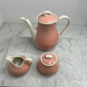 Op de afbeelding: Een vintage theeservies in een zachte roze tint met crèmekleurige accenten. De set bevat een theepot met deksel, een melkkan en een suikerpot met deksel. De theepot heeft een gebogen tuit en handvat. De set is waarschijnlijk gemaakt van keramiek.