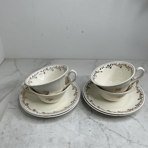 Könnte beinhalten: Vier cremefarbene Tassen und Untertassen. Jede Tasse und Untertasse hat ein goldenes Blumendesign am Rand und am Boden der Tasse. Die Tassen haben einen gebogenen Griff. Die Untertassen sind rund.