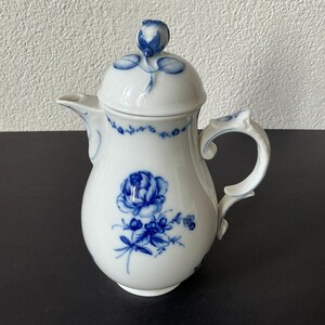 Può includere: Una caffettiera in porcellana bianca con un motivo floreale blu. La caffettiera presenta un manico curvo, un beccuccio e un coperchio sormontato da una rosa blu. Il corpo è decorato con una grande rosa blu e piccoli accenti floreali.