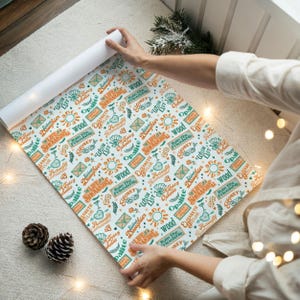 May include: A roll of patterned wrapping paper with a white background. The design features various words and phrases in teal and orange, including "The Life of a Showgirl" and "Elizabeth Taylor". The paper is being unrolled.