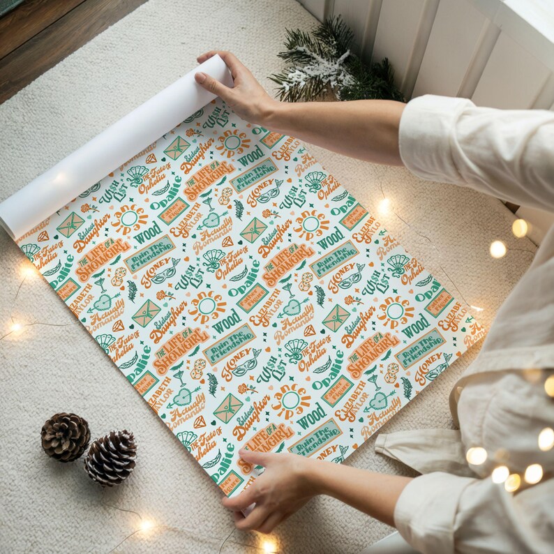 May include: A roll of patterned wrapping paper with a white background. The design features various words and phrases in teal and orange, including "The Life of a Showgirl" and "Elizabeth Taylor". The paper is being unrolled.