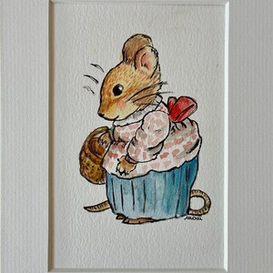 May include: Watercolor painting of a mouse dressed as a cupcake, holding a basket. The mouse has brown fur, a pink and white patterned top, and a blue cupcake base. A red bow is on the back. The artwork is framed in white.