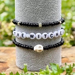 May include: Three black beaded bracelets stacked on a gray surface. The top bracelet has silver beads. The middle bracelet has white beads that spell out "THE WITCH." The bottom bracelet has a white skull charm.