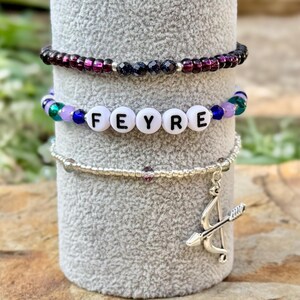 May include: Three beaded bracelets displayed on a gray cylinder. The top bracelet is black and purple. The middle bracelet spells "FEYRE" with white letter beads. The bottom bracelet has a silver bow and arrow charm.