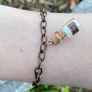 May include: A brown chain bracelet with a small glass bottle charm. The bottle is filled with glitter and has a cork stopper. The bottle has a label that says "Wish dust".