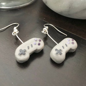 Puede incluir: Un par de pendientes grises con forma de mandos de videojuegos retro. Los mandos tienen detalles de botones morados y grises y están suspendidos de ganchos plateados. Los pendientes se muestran sobre una superficie marrón oscuro.