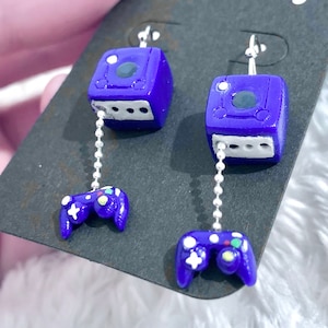 May include: A pair of handmade purple GameCube earrings. Each earring features a miniature GameCube charm with a matching controller dangling below. The controllers have colorful button details and are connected to the GameCube charms by a silver chain.