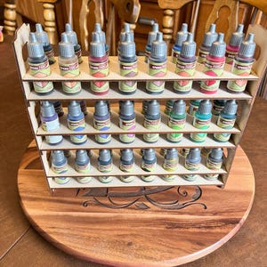 Puede incluir: Un organizador de botellas de pintura de madera con múltiples estantes, lleno de pequeñas botellas de Speed Paint en varios colores. El organizador se encuentra sobre una base redonda de madera con tallas decorativas.