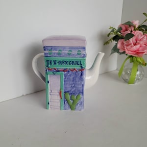 May include: A white ceramic teapot shaped like a Tex-Mex restaurant building with a green door, red chili peppers, and a cactus. The teapot has a purple roof and blue walls with the words "Tex-Mex Grill" written in green.