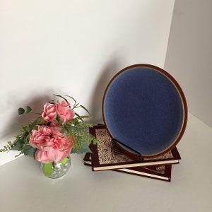 May include: A blue ceramic plate with a brown rim sits on top of a stack of three brown books with gold accents. A vase with pink flowers sits in the foreground.