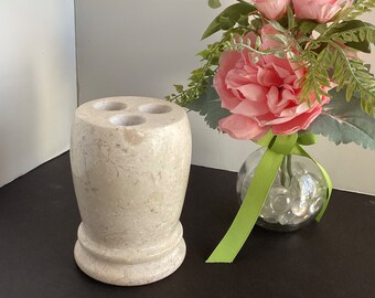 May include: A white marble toothbrush holder with three holes. The holder has a textured surface and is sitting on a black surface. A vase with pink flowers and green leaves is in the background.