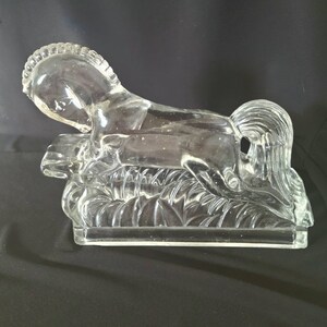 May include: A clear glass sculpture of a horse lying down on a bed of leaves. The horse is detailed with a mane and tail.